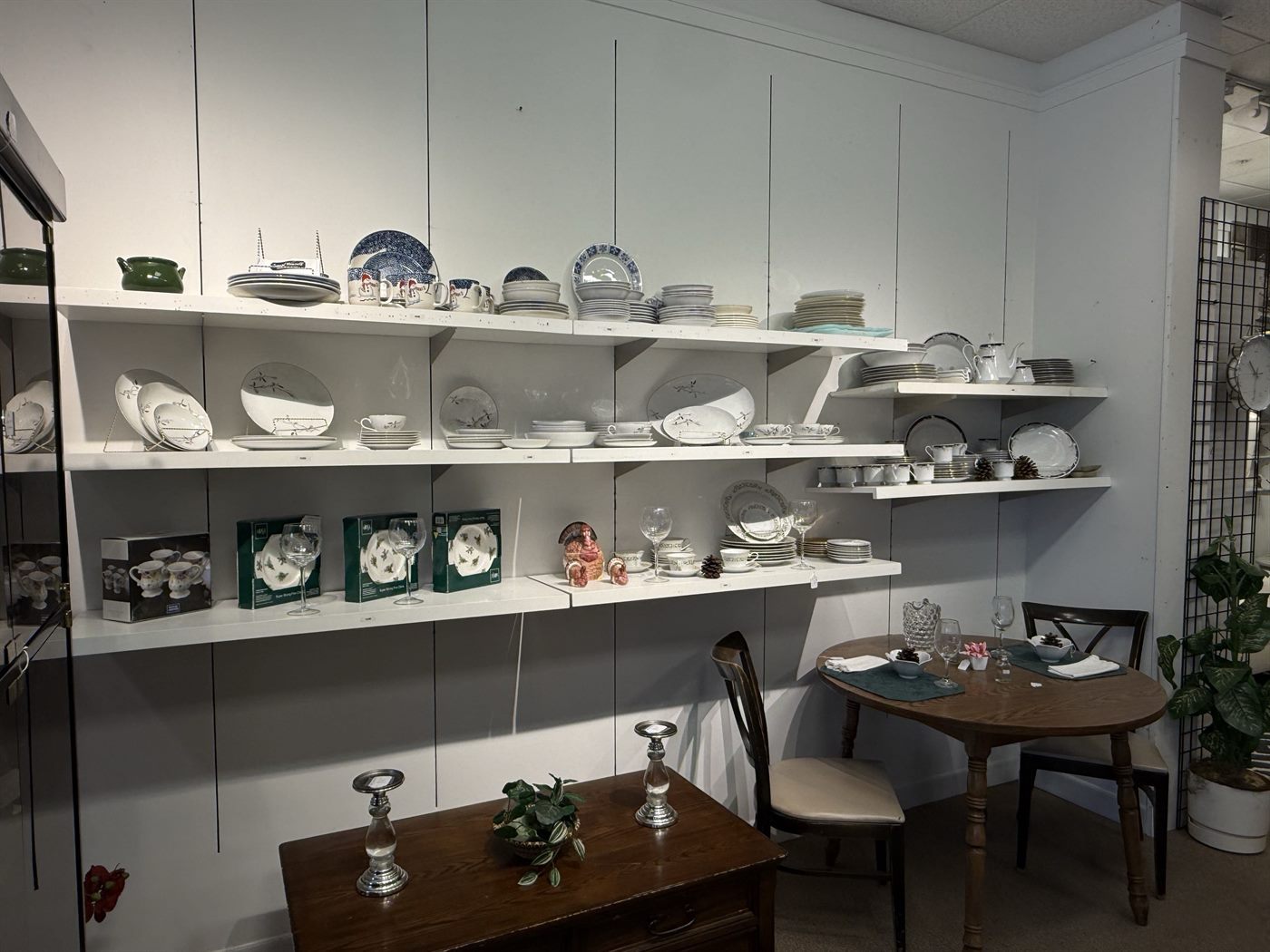 Wall shelves of fine china, dinnerware sets and stemware above a set dining table at K&A Thrift